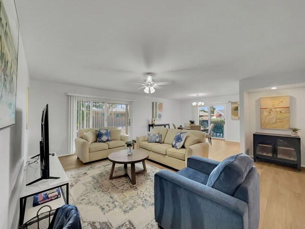 Ein modernes Wohnzimmer mit einem beigen Sofa, einem blauen Sessel und einem Couchtisch. Ein Fernseher steht auf einem Lowboard, es gibt einen Deckenventilator und ein Bild an der Wand.