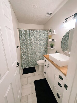 A bathroom with a shower curtain, toilet, sink, mirror, and shelves with toiletries.