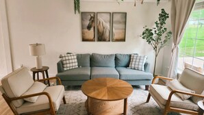 Una sala de estar con un sofá, dos sillones, una mesa de centro y una lámpara. Hay tres cuadros enmarcados con motivos ecuestres en la pared, una planta en el alféizar de la ventana y vistas a un césped verde al exterior.
