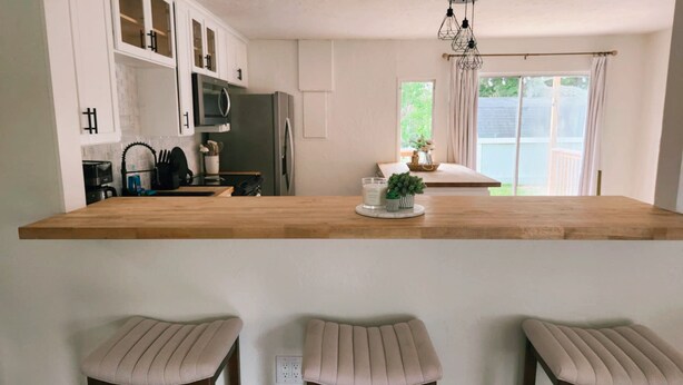 Una cocina con armarios blancos, encimera de madera y tres taburetes de bar.