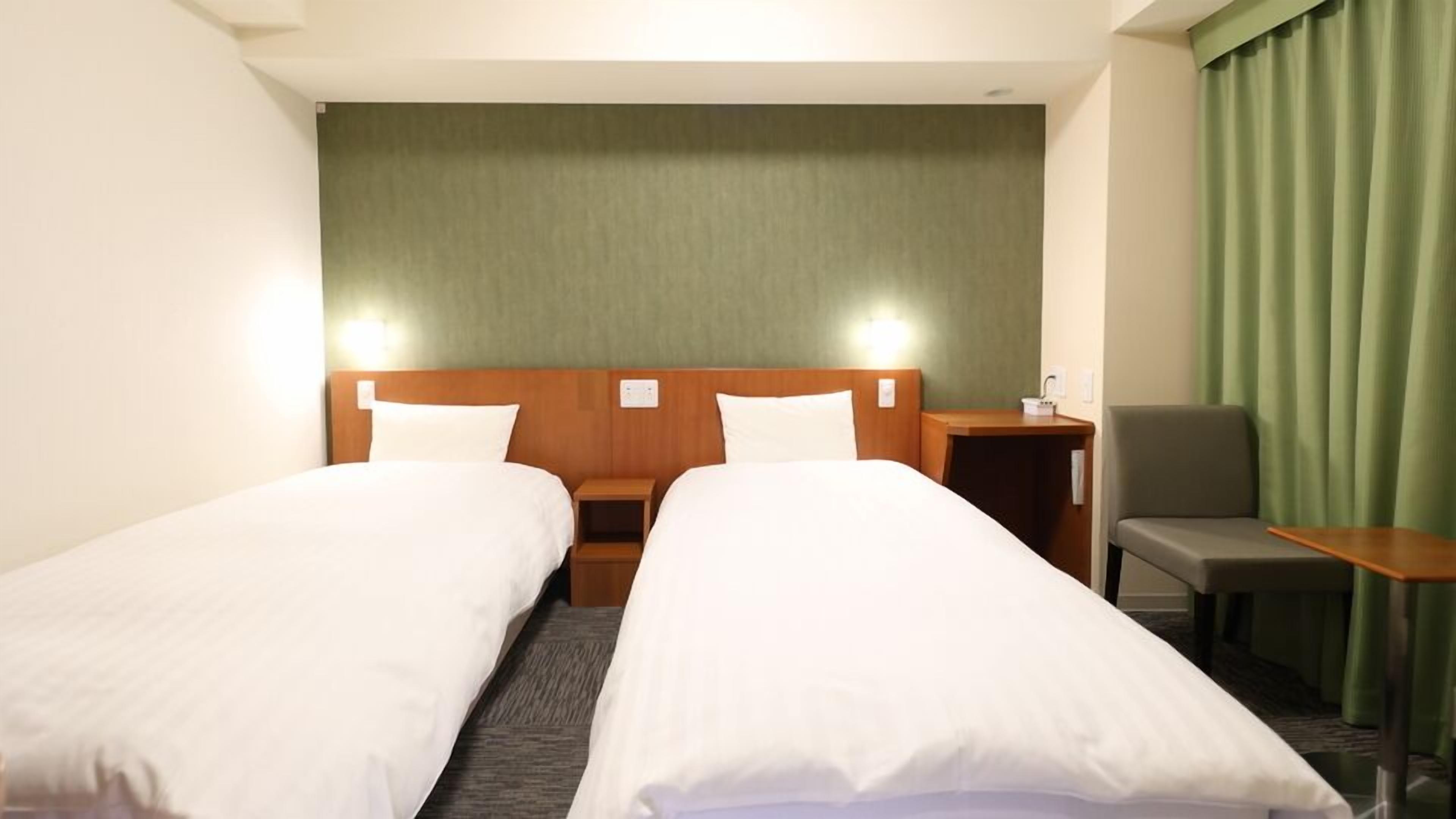 A hotel room with two single beds, a small table, a chair, and a green curtain.