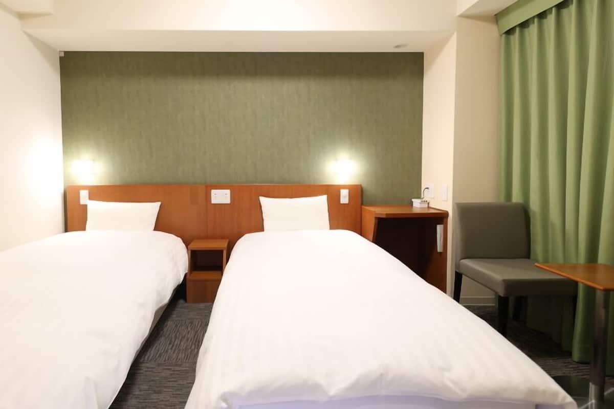 A hotel room with two single beds, a small table, a chair, and a green curtain.