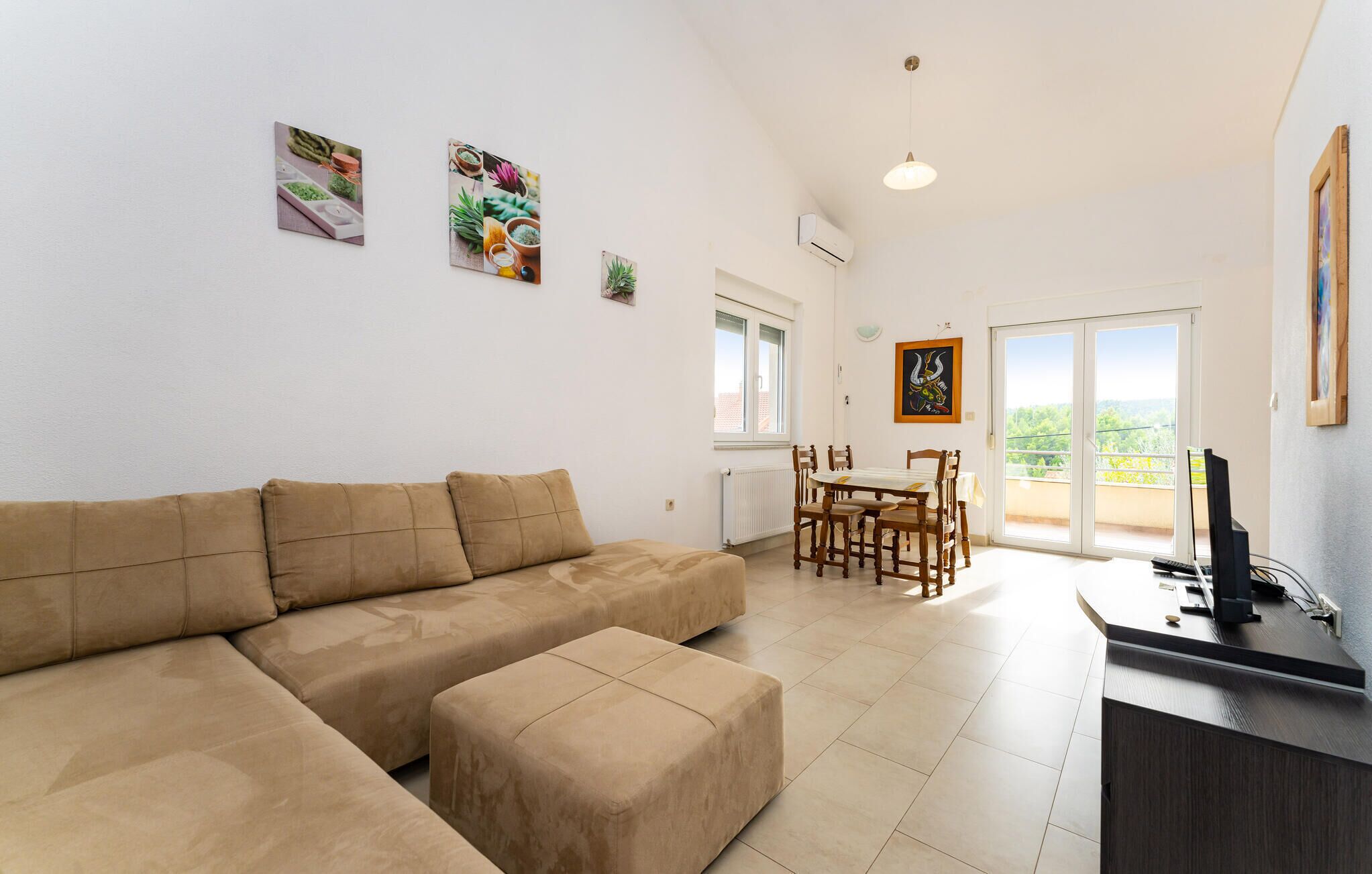 A living room with a beige sofa, a dining table with chairs, and a balcony with a view.