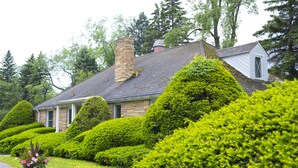 Une maison en pierre avec un toit à pignon et une cheminée, entourée de haies et d’arbres bien entretenus.
