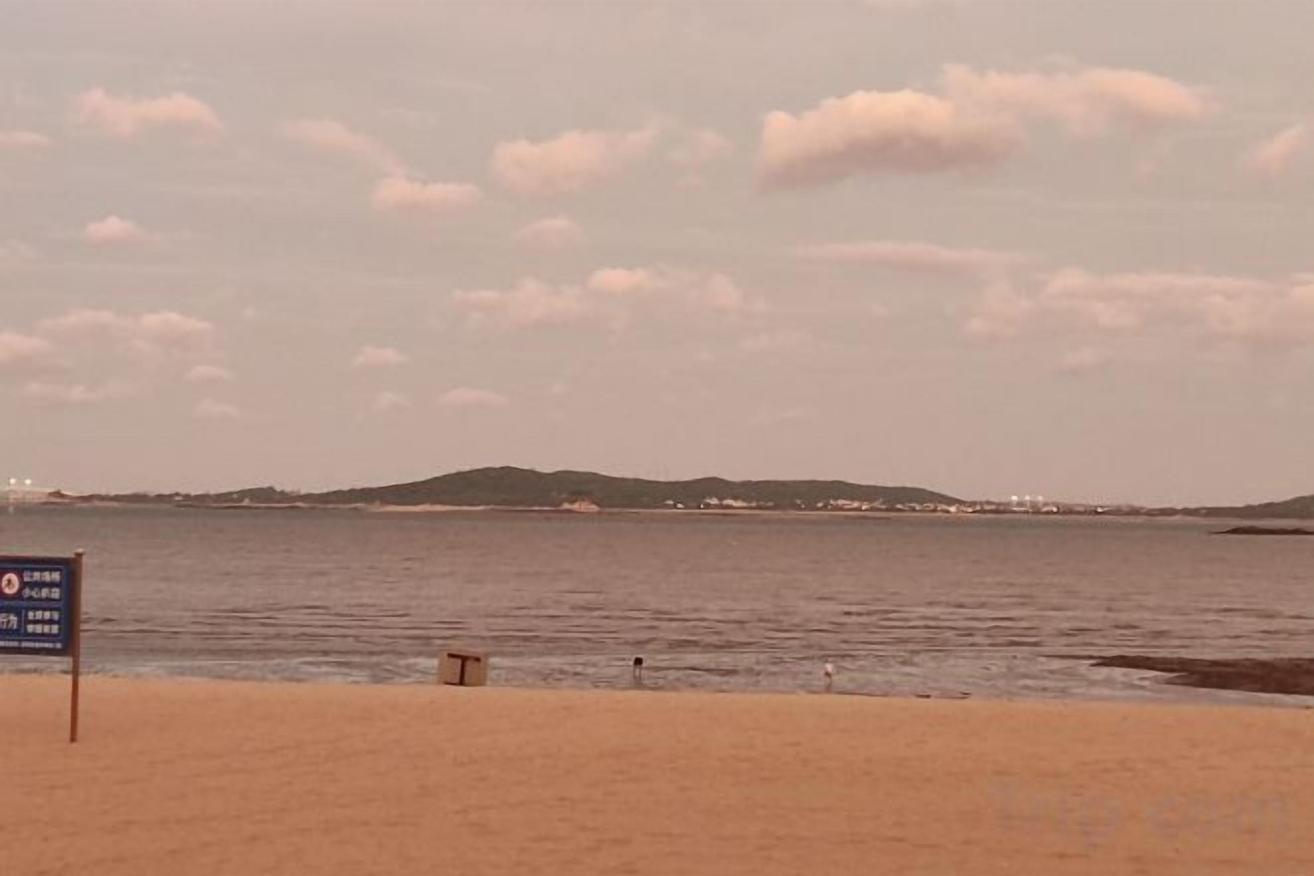 A beach with a sign in the foreground, a calm sea, and a distant shoreline with hills.