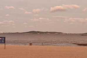 A beach with a sign in the foreground, a calm sea, and a distant shoreline with hills.
