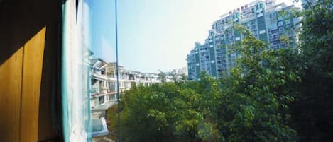 A view through a window with a building in the background, a balcony, and some greenery.