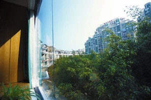 Vista a través de una ventana con un edificio al fondo, un balcón y algo de vegetación.