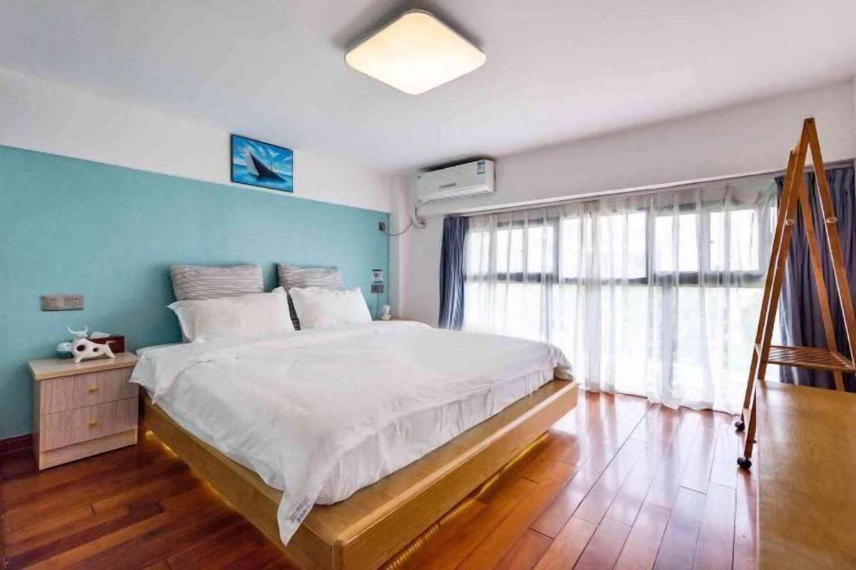 Une chambre avec un grand lit, un plancher en bois, une table de chevet, un climatiseur fixé au mur et une fenêtre avec des rideaux.
