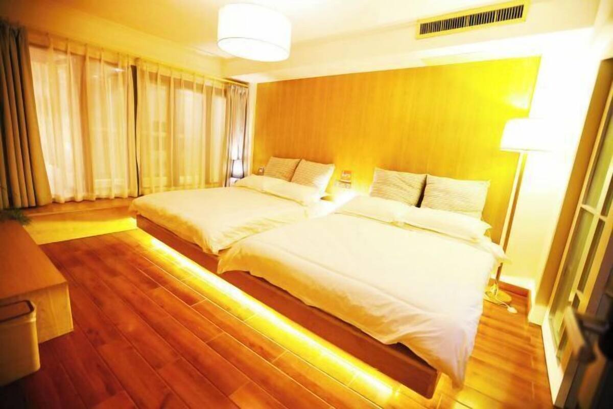 Une chambre d’hôtel avec deux lits, un plancher en bois, une grande fenêtre avec des rideaux et une bouche d’aération au plafond.