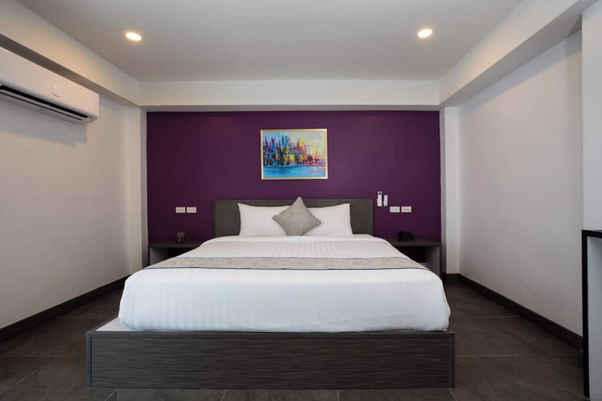 Una habitación de hotel moderna con una cama grande, paredes de acento moradas y un cuadro de paisaje urbano.