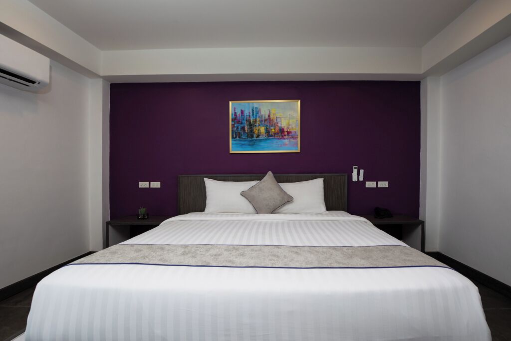 A hotel room with a large bed, purple accent walls, a cityscape painting, and a small bedside table with a lamp.