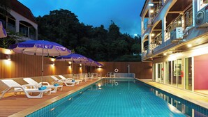 Una piscina con sillas de descanso y sombrillas, rodeada por una terraza de madera y un edificio con balcones.