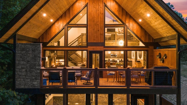 Una cabaña de madera grande con una terraza amplia, una chimenea de piedra y una cocina visible a través de ventanas grandes.