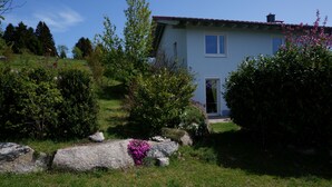 A house with a light blue exterior, a red roof, and a front door. There are green bushes and a rock with pink flowers in front of the house. - Quiet vacation apartment in the southern Black Forest, 10 minutes from St. Blasien (Dachsberg (Südschwarzwald))
