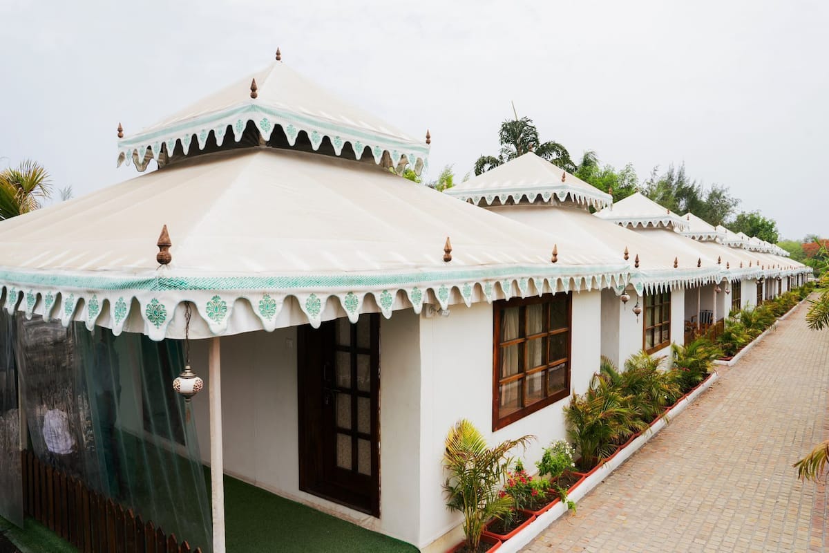 A row of white tents with green trim and decorative elements, lined with potted plants along a paved walkway.