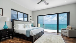 A bedroom with a large bed, a nightstand, a lamp, a chair, and a view of the ocean through large windows. - 3 BR Oceanfront Beach House with Private Pool Gated Community Staircase to Ocean (Nassau)
