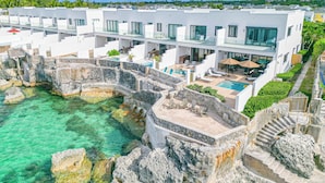A modern resort with a pool surrounded by stone walls, a covered outdoor area with lounge chairs, and a view of the ocean. - 3 BR Oceanfront Beach House with Private Pool Gated Community Staircase to Ocean (Nassau)