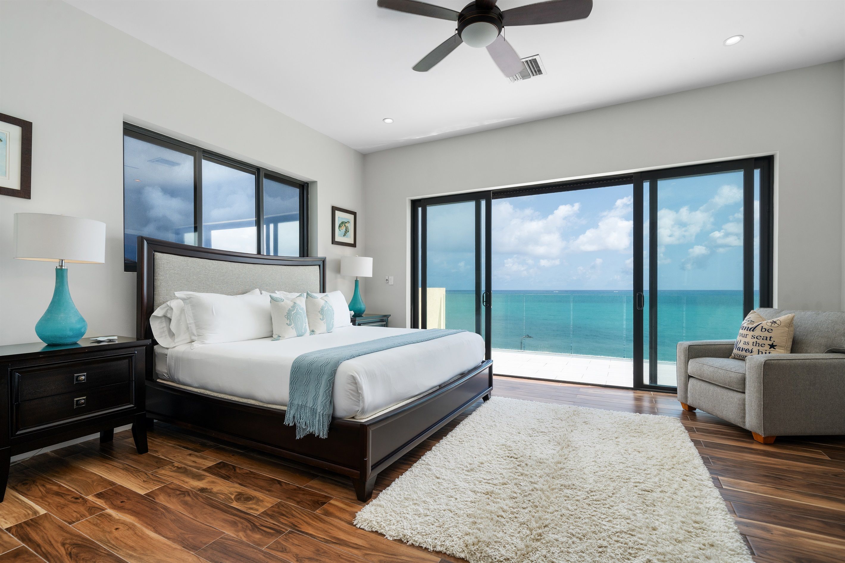 A bedroom with a large bed, a ceiling fan, a nightstand, a lamp, a chair, and a view of the ocean through large windows.