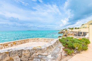 A coastal view with a stone wall, a small garden, and a building with a balcony overlooking the sea. - Oceanfront with Beach Access & Private Pool 3BR (Nassau)