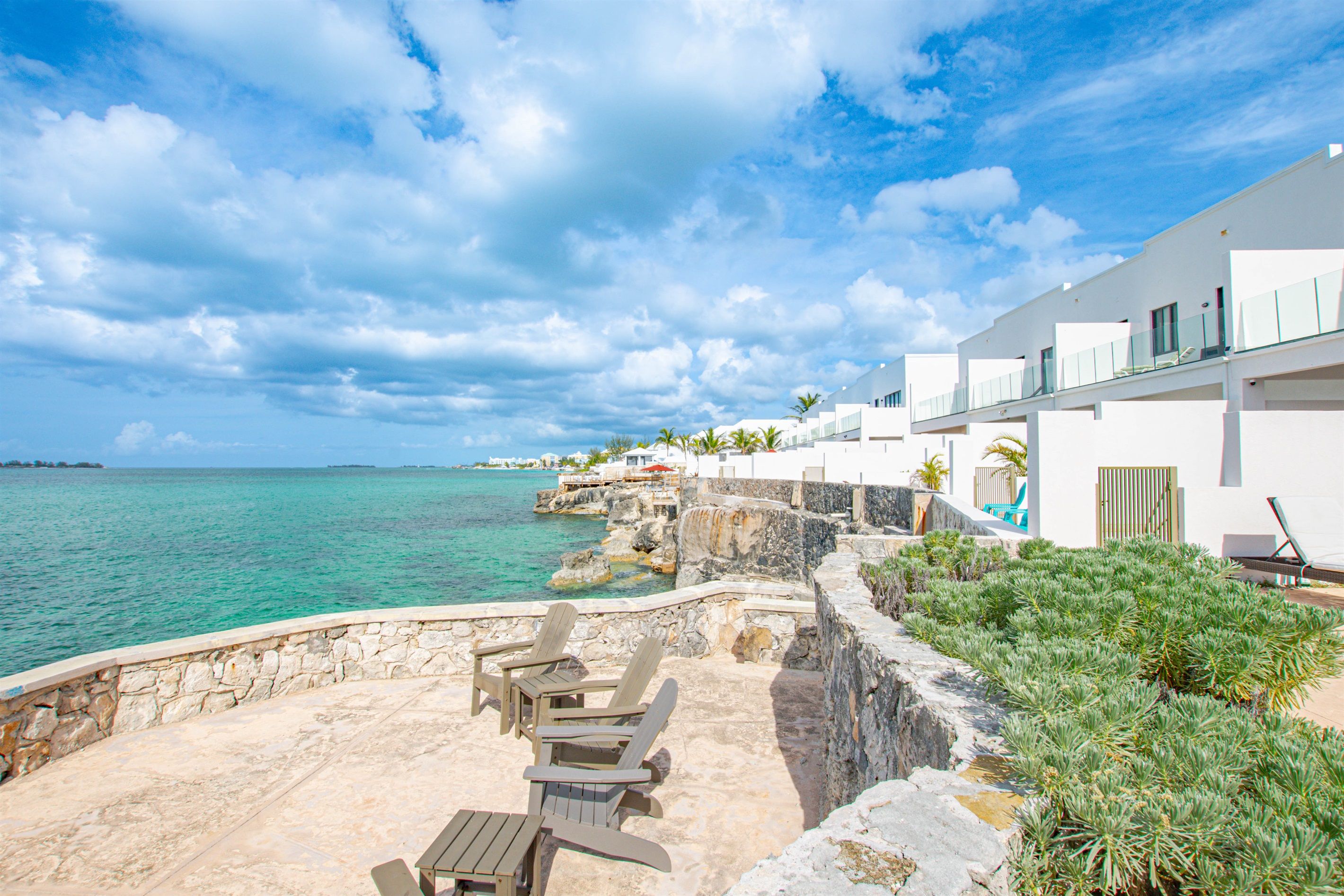 A coastal hotel with a stone patio, outdoor seating, and a view of the sea and distant island.