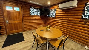 Ein gemütliches Blockhauszimmer mit einem Holzesstisch, vier Stühlen, einer Fernbedienung und einer Vase mit Sonnenblumen.