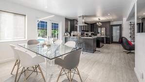 Une cuisine moderne avec un îlot central, des chaises blanches disposées autour d’une table en verre, et une vue sur la verdure à l’extérieur.