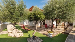 Une cour avec un sol en pierre, de l’herbe verte et des arbres. On y trouve des espaces de détente en plein air, avec des chaises et des tables rouges.