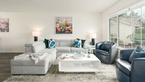 Ein modernes Wohnzimmer mit einem grauen Ecksofa, einem weißen Couchtisch und einem schwarzen Ledersessel. Zwei Stehlampen und ein großes Fenster sorgen für natürliches Licht.