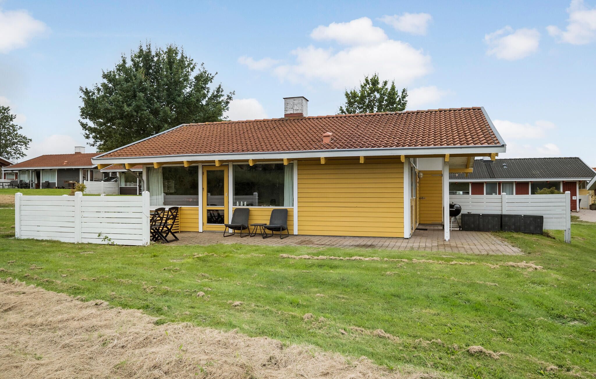 Een geel gebouw met een pannendak, een overdekte patio met stoelen, en een omheind gazon.