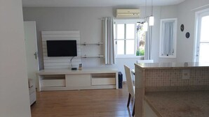 A modern living room with a flat-screen TV mounted on a white wall, a built-in entertainment unit, a dining area with a kitchen counter, and a window with curtains. - Pousada Anambé (Angra dos Reis)