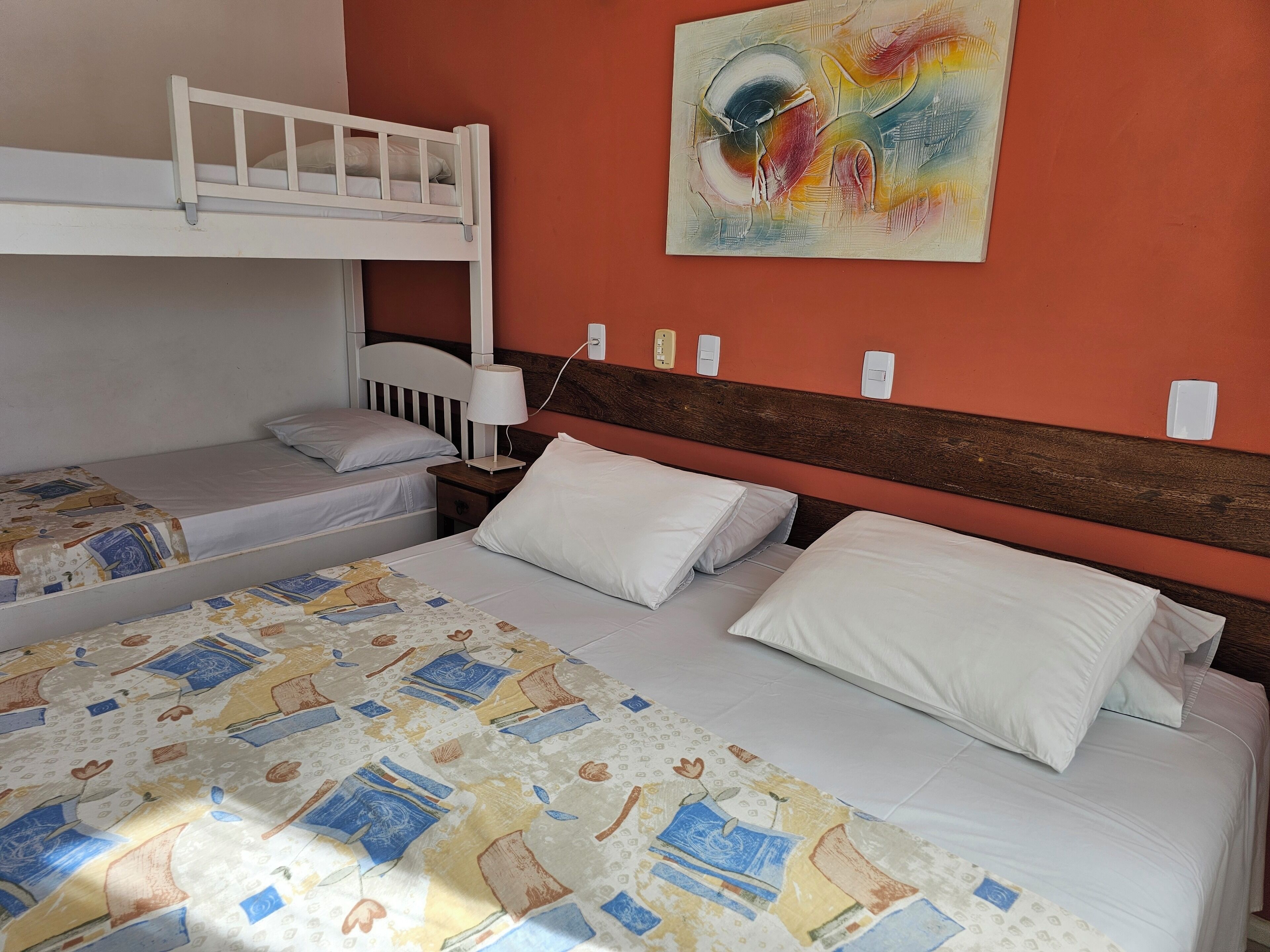 Habitación con dos camas, una litera y otra individual. Hay una mesita de noche con una lámpara y un enchufe. Un cuadro abstracto y colorido adorna la pared.
