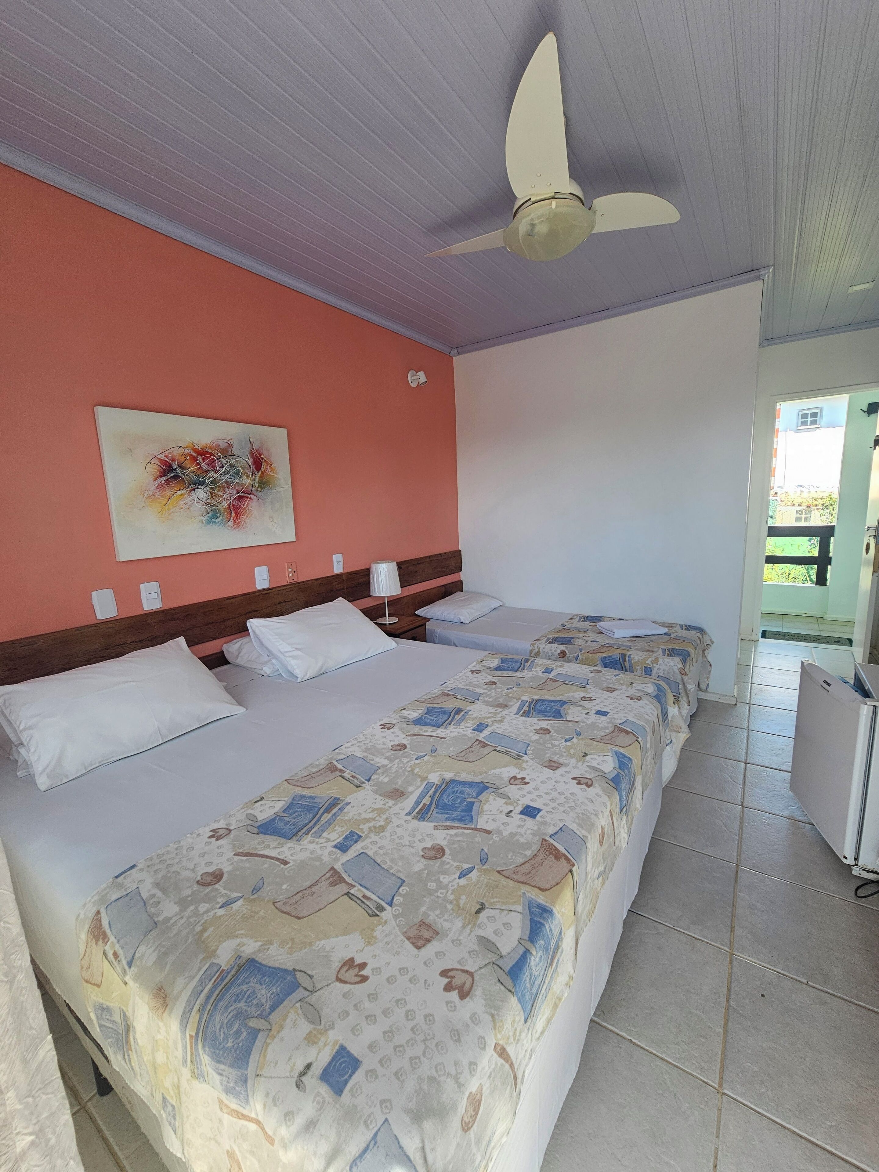 Ein Hotelzimmer mit zwei Betten, einem Deckenventilator, einem Bild an der Wand und Blick nach draußen.
