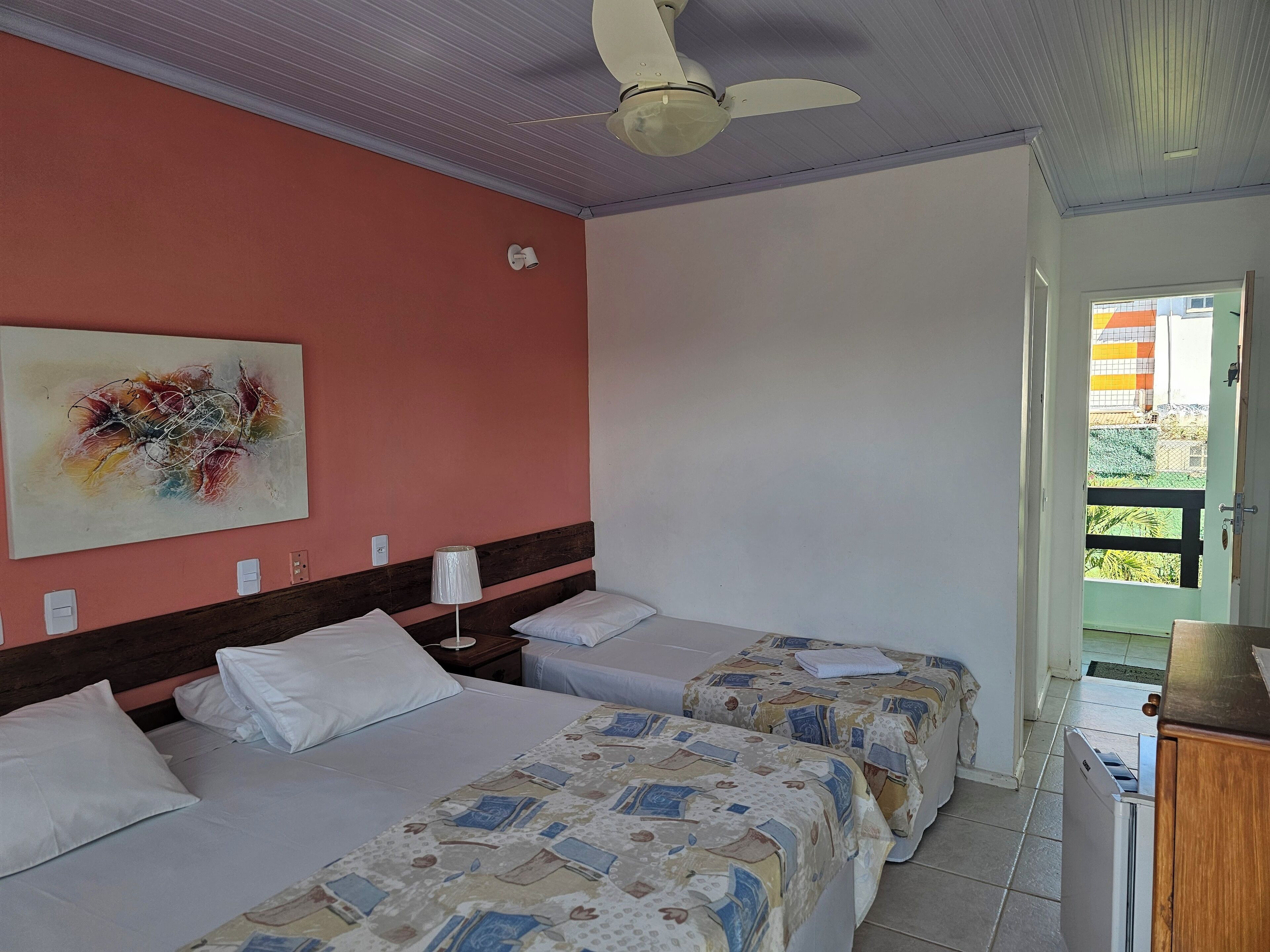 A hotel room with two beds, a ceiling fan, a painting on the wall, and a view of the outside.