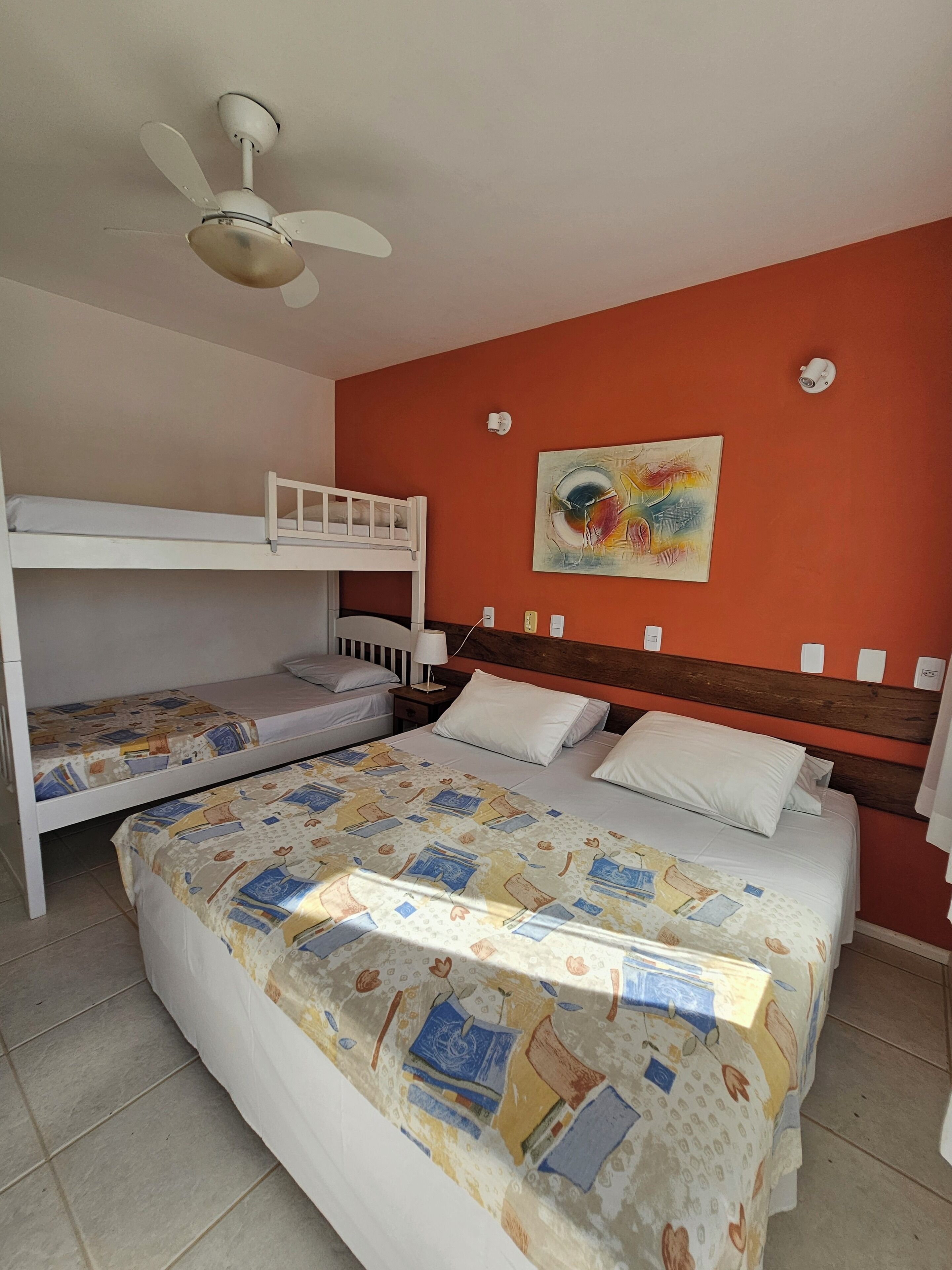 A bedroom with a bunk bed, a single bed, a ceiling fan, a painting on the wall, and a clock.