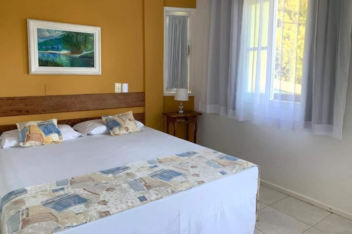 A bedroom with a large bed, a painting on the wall, a window with curtains, and a nightstand with a lamp.