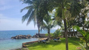 A coastal garden with a stone pathway, palm trees, and lounge chairs by the water. - Charming 1-bedroom in Tower Isle St Mary: AC, WiFi, pool, and a serene retreat. (Tower Isle)