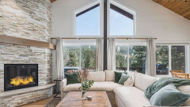 A living room with a stone fireplace, a large sectional sofa, a wooden coffee table, and a view of a forest through large windows. - Renovated Ski Home with a Hot Tub & Views of Slope - Perfect Ski Lease (Incline Village)