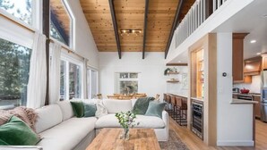 A modern living room with a wooden ceiling, white sofas, a coffee table, and a kitchen area visible through an open doorway. - Renovated Ski Home with a Hot Tub & Views of Slope - Perfect Ski Lease (Incline Village)