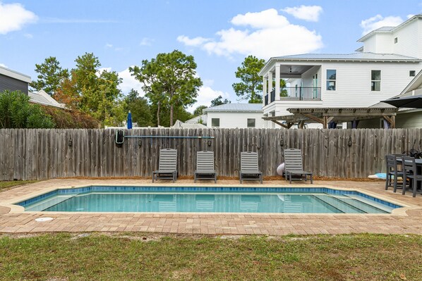 A rectangular swimming pool with a tiled edge, surrounded by a brick patio. There are four lounge chairs aligned against a wooden fence.