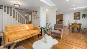 Un salon moderne avec un plancher en bois, un canapé jaune, une table basse blanche et un fauteuil gris. Un escalier avec des rampes blanches mène à un autre niveau.