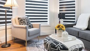 Een moderne woonkamer met een grijze bank, een grijze fauteuil, een ottoman met een patroon, een vloerlamp en een vaas met zonnebloemen.