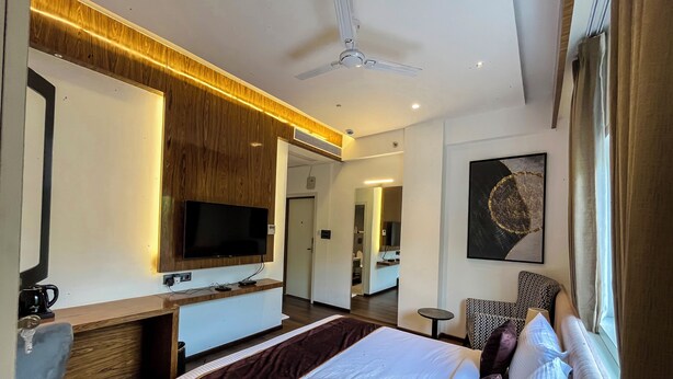 A hotel room with a bed, a desk, a TV, a ceiling fan, and a large window. - Wishotel Inn Rasa Royal - A Boutique Hotel (Indore)