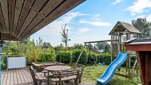 Une terrasse en bois avec une table de pique-nique et des bancs, un toboggan bleu et une cabane de jeux en bois.