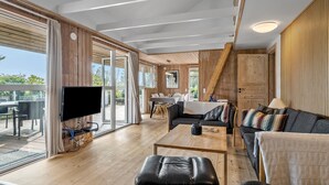 Un vaste espace de vie avec une grande porte-fenêtre coulissante donnant sur une terrasse extérieure, un téléviseur à écran plat fixé au mur, un canapé, une table basse et un plafond en bois avec des poutres apparentes.