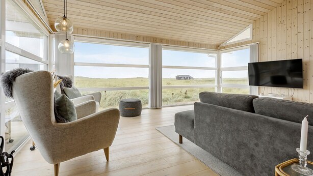 A modern living room with a large window, a grey sofa, a chair with a fur throw, and a flat-screen TV mounted on the wall. - 3 bedroom ocean front home in Hjørring (Hjørring)