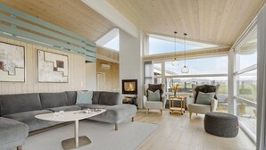 A modern living room with a grey sectional sofa, a round coffee table, and a fireplace. There are two armchairs and a round ottoman. The room has wooden floors, a large window, and a view of the outdoors. - 3 bedroom ocean front home in Hjørring (Hjørring)