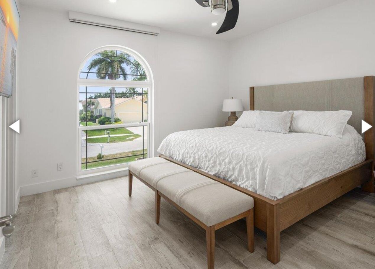 A bedroom with a large bed, a bench, a window with a view of a house and palm trees, and a ceiling fan.
