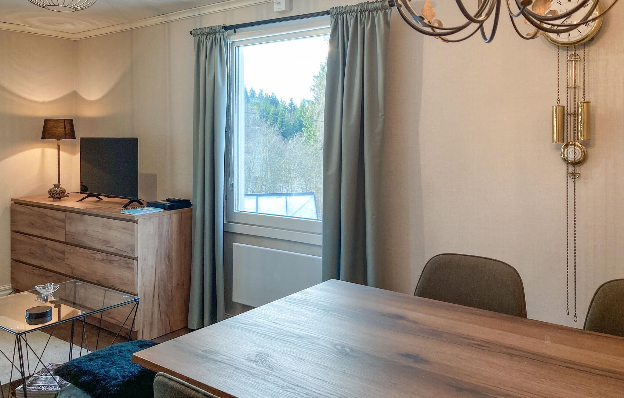 A room with a wooden desk, a television, a dresser, a glass table, and a window with curtains.