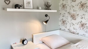 A bedroom with a white nightstand, a bed with a white headboard, a wall-mounted shelf with decorative items, a floral wallpaper, and a bedside lamp. - Nice home in Sävsjö with WiFi (Sävsjö)